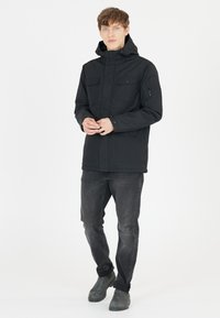 Sort vandtæt jakke med hætte, der har to brystlommer og en lynlåslomme på ærmet. Parret med mørke jeans og grå støvler.