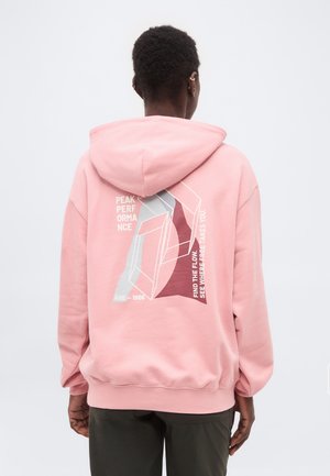 Άτομο που φοράει ροζ φούτερ με κουκούλα με αφηρημένο γεωμετρικό σχέδιο και κείμενο που γράφει «Peak Performance» και «Find the flow, see where free takes you» στην πλάτη.