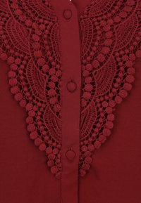 Burgundská blůza s propracovanými krajkovými detaily a texturovanými bobble akcenty. Obsahuje centrální zapínání na knoflíky a měkkou látku.