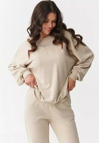Beżowa bluza i pasujące spodnie, oba wykonane z miękkiego materiału. Bluza ma wyraźny, wytłaczany nadruk na klatce piersiowej oraz luźny krój.