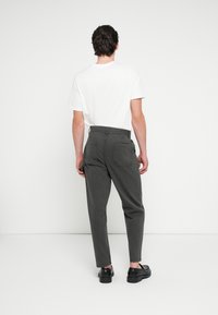 Graue Baumwollhosen mit schmal zulaufender Passform, ausgestattet mit zwei Gesäßtaschen und einer Knopfverschluss. Kombiniert mit einem weißen T-Shirt und schwarzen Schuhen.