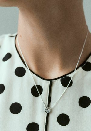 Vrat nosi srebrnu ogrlicu s okruglim privjeskom preko bijele bluze s velikim crnim točkicama i crnim rubom.