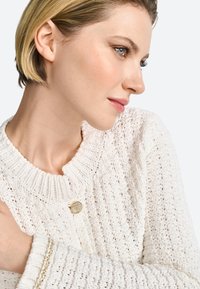 Cardigan en tricot blanc avec un motif texturé, un col rond et un détail de bouton doré à l'avant. Fil doux et coupe décontractée.