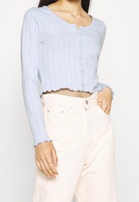 Svetle modrý zkrácený cardigan, žebrovaná textura, zaoblený výstřih s knoflíkovým zapínáním, vlnkový lem a manžety, v kombinaci se světle růžovými kalhotami.