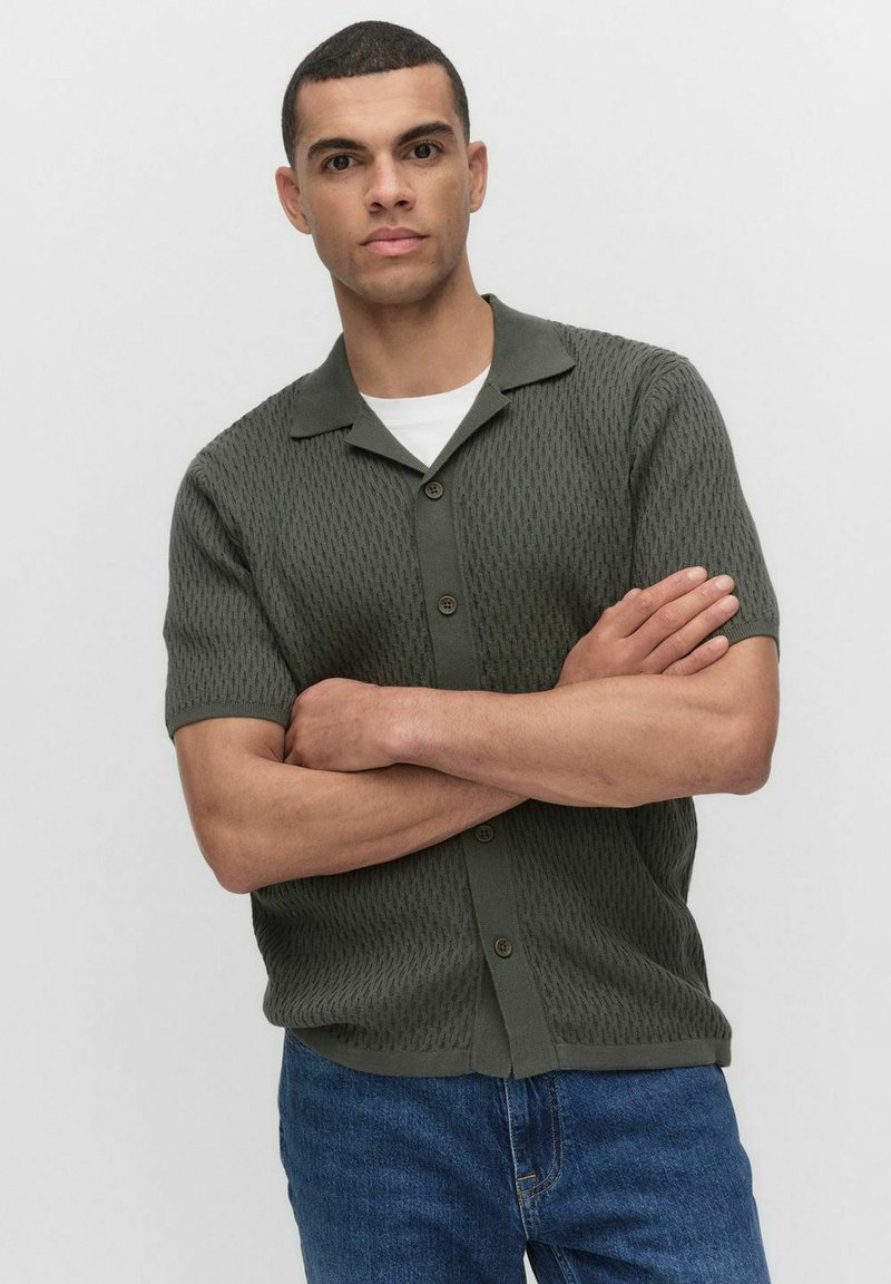 Korte mouwen, donkergroen gebreid shirt met een textuurpatroon, knoopsluiting en een scherpen kraag, gecombineerd met blauwe jeans.