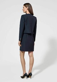 Granatowa marynarka o skróconym kroju, zestawiona z dopasowaną spódnicą. Złote guziki na rękawach, gładki materiał i czarne, szpiczaste obcasy.