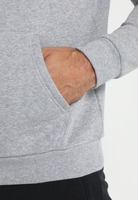 Sudadera gris con un bolsillo canguro y puños acanalados. El material parece suave con una textura lisa, mostrando un diseño casual.