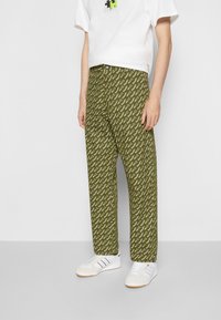 Pantalon vert à motifs avec un design répétitif de logo blanc, présentant une coupe décontractée, associé à un t-shirt blanc et des baskets blanches.
