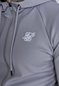 Sivi dukserica sa patentnim zatvaračem izrađena od glatkog materijala, sa bijelim izvezenim logotipom na prsima i podešavanjima na kapuljači.