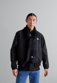 Jacketa bomber neagră cu manșete elastice, combinată cu o eșarfă lungă din blană artificială neagră. Modelul poartă blugi albaștri și un top negru dedesubt.