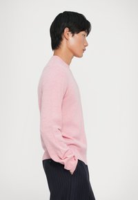 Svijetlo ružičasti pulover s okruglim izrezom, mekanog teksture i dugih rukava, u kombinaciji s tamnim hlačama s prugicama. Jednostavan, usko rezan dizajn.