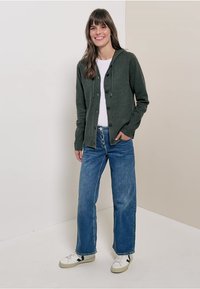 Ciemnozielony sweter z dzianiny z zapięciem na guziki z przodu i kapturem, w połączeniu z białą koszulą oraz niebieskimi spodniami typu wide-leg. Całość dopełniają białe sneakersy.