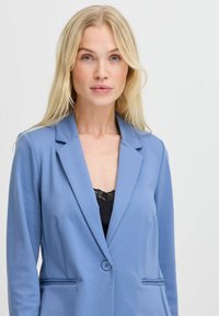 Blazer bleu clair en tissu lisse, avec un col cranté, fermeture à un bouton et poches latérales. Accentué par une bordure en dentelle en dessous.