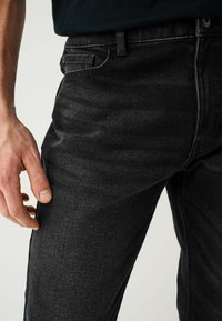 Jeanși negri din denim expuși, având o croială dreaptă, textură subtilă, cusături în contrast și buzunare laterale. O mână sprijinită pe material.