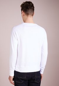 Vitt långärmat sweatshirt med rund hals, ribbade muddar och rak nederkant, kombinerad med mörkblå jeans i en avslappnad miljö.