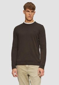 Brauner Rundhalsausschnitt-Pullover mit ribbedem Saum und Bündchen, mit weißem Innenkragen. Getragen über beigen Hosen, minimale Markenkennzeichnung sichtbar.