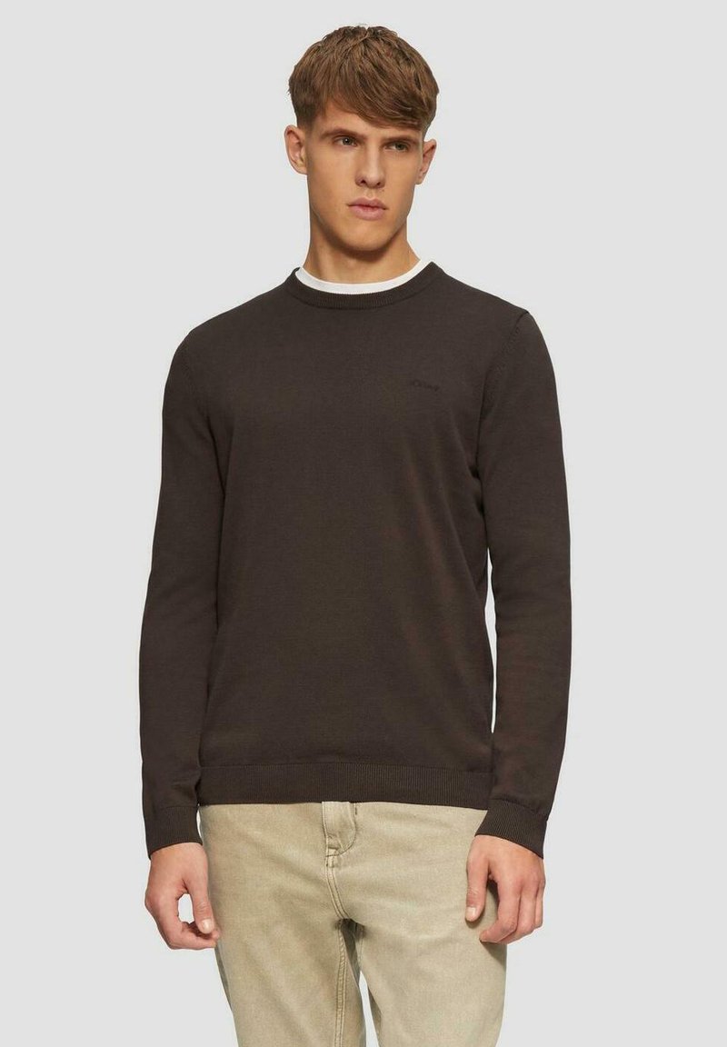 Brauner Rundhalsausschnitt-Pullover mit ribbedem Saum und Bündchen, mit weißem Innenkragen. Getragen über beigen Hosen, minimale Markenkennzeichnung sichtbar.
