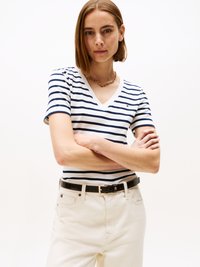 T-shirt riscado de decote em V em azul marinho e branco, combinado com calças de cor creme e um cinto preto. A camisa tem um pequeno detalhe de logótipo no peito.