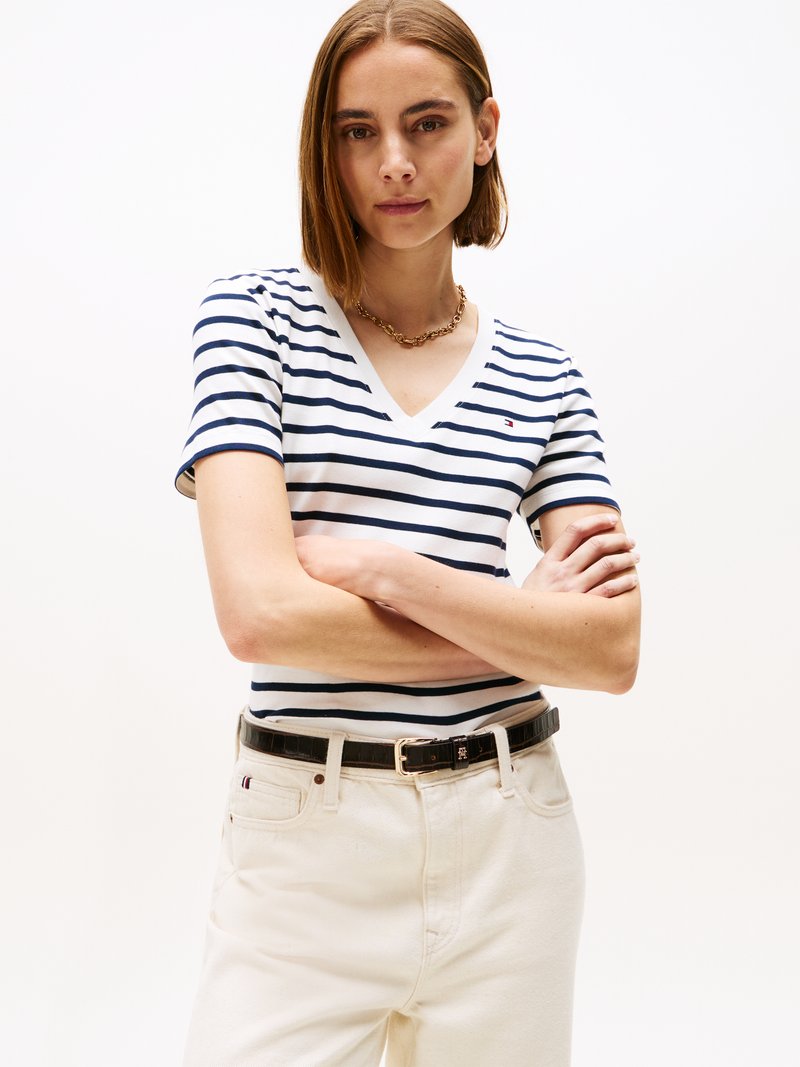 T-shirt riscado de decote em V em azul marinho e branco, combinado com calças de cor creme e um cinto preto. A camisa tem um pequeno detalhe de logótipo no peito.