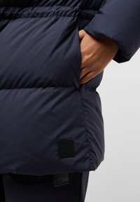 Marine blauwe pufferjack met een gladde, glanzende afwerking. Voorzien van een zijzak en een zwart logo patch op de zoom. Bevat gerimpelde details in de taille.