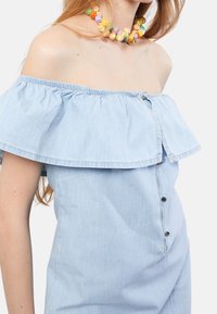 Vaaleansininen olkaimeton farkkupaita, jossa on röyhelöreunus ja etupainonapit. Sileä rakenne elastisilla reunoilla.