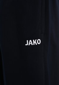 Schwarze Sporthose mit einem weißen gestickten "JAKO"-Logo am linken Bein. Das Material wirkt glatt mit einer matten Textur.