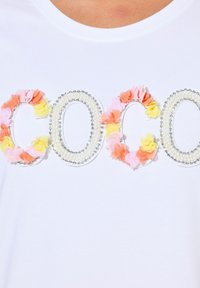 Camiseta blanca con la palabra "COCO" decorada con aplicaciones de flores en tonos pastel y acentos de cuentas, creando un diseño texturizado.