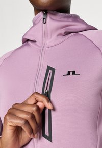 Sudadera con cremallera de color púrpura claro hecha de tela suave, con un bolsillo de cremallera negra y acento de logo. Primer plano de la cremallera y la interacción de la mano.