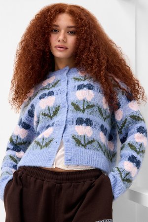 Gilet en tricot de couleur bleu clair avec des motifs floraux en rose, marine et vert. Présente une finition texturée et une fermeture à boutons.