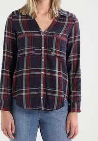 Marinegekleurde geruite blouse met rode en grijze lijnen, knoopsluiting aan de voorkant, twee borstzakken, lange mouwen en een kraag. Zachte stof met een ontspannen pasvorm.