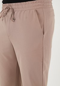 Lichtbruine broek met elastische tailleband en zijzakken, van een gladde stof en met een ontspannen pasvorm. De trekkoorden met metalen uiteinden zijn zichtbaar.