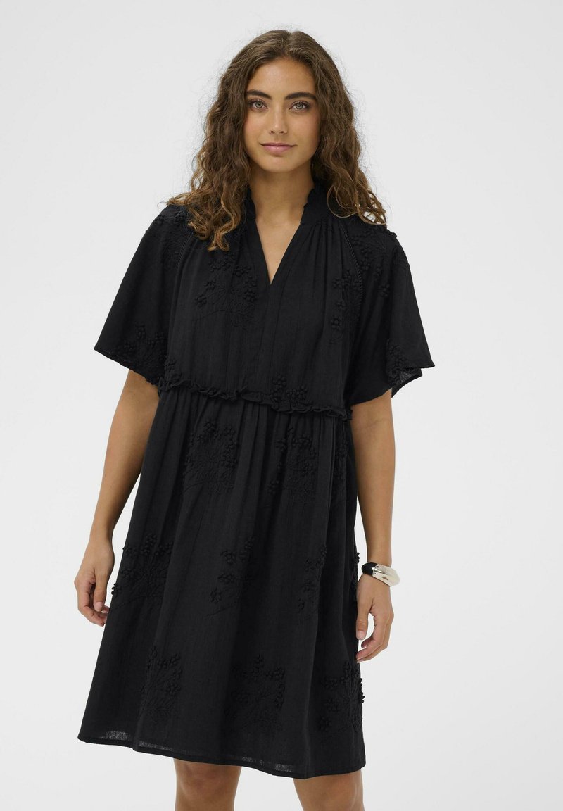 Femme aux longs cheveux bouclés portant une robe noire à manches courtes avec une broderie florale et un bracelet blanc au poignet droit.