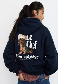 Donna che indossa una felpa con cappuccio blu navy con una grafica di un bassotto con cappello da chef e testo "Le Chef Bon appétit Made with paws and love."
