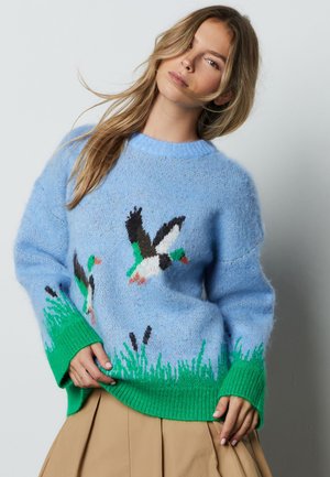 Woman with long hair wearing a blue sweater featuring flying ducks and green grass, paired with a beige pleated skirt.