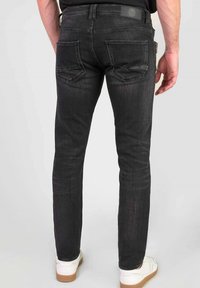 Hombre vestido con pantalones vaqueros ajustados negros y zapatillas blancas, de pie con la espalda hacia la cámara frente a un fondo liso.