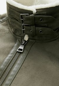 Chaqueta de ante verde oliva con un cuello forrado de lana, que presenta un cierre frontal con cremallera y dos correas de cuero con acentos de metal.