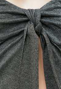 Κοντινό πλάνο από ένα υφασμάτινο υλικό σε ασημένιο-γκρι χρώμα με υφή, δεμένο σε έναν μεγάλο κόμπο, αποκαλύπτοντας μια μικρή σχισμή δέρματος από κάτω.