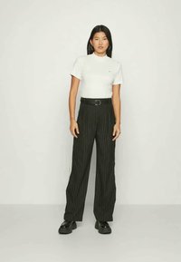 Mulher de pé, vestindo uma blusa branca de manga curta, calças largas pretas com riscas verticais e um cinto, e sapatos em plataforma pretos, em frente a uma parede simples.