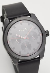 Μαύρο ρολόι με δερμάτινο λουρί που διαθέτει σκούρο γκρι καντράν, κόκκινες λεπτομέρειες, υποδείκτες ημερομηνίας και ημέρας, και μινιμαλιστικό σχεδιασμό με έντονα ωρολογιακά δείκτες.