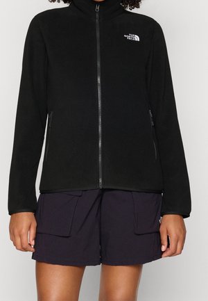 Person iført en sort fleecetrøje med lynlås med The North Face-logo og sorte cargoshorts mod en ensfarvet baggrund.