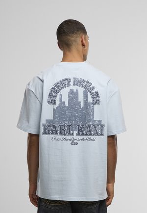 T-shirt oversize bleu clair avec un imprimé graphique représentant une ligne d'horizon de la ville et le texte "STREET DREAMS" et "KARL KANI" au dos. Manches courtes.