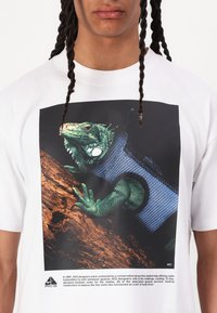 Tricou alb din bumbac, cu un grafic mare al unei iguane verzi cu accente de material albastru, pe un fundal întunecat cu detalii texturate.