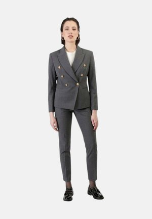 Donna in piedi che indossa un blazer doppiopetto grigio con bottoni dorati, pantaloni slim abbinati, scarpe nere in vernice e orecchini a cerchio.