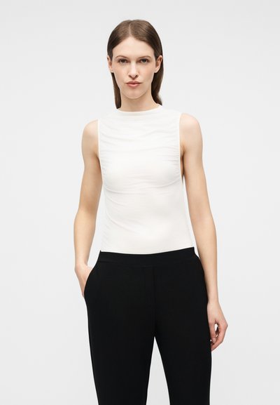 Γυναίκα με ίσια καστανά μαλλιά, φορώντας λευκό τοπ χωρίς μανίκια και μαύρο παντελόνι, στέκεται με το ένα χέρι στην τσέπη, μπροστά σε λευκό φόντο.