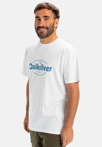 Bílý bavlněný tričko s krátkým rukávem a kulatým výstřihem. Na přední straně je modrý grafický logo se textem "Quiksilver".