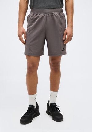 Mann trägt graue Sportshorts, weiße Markensocken und schwarze Sportschuhe, steht vor einem einfarbigen Hintergrund.