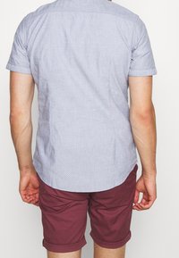 Světle modrá košile s krátkým rukávem, s tečkovaným vzorem, límečkem a zakulaceným lemem, spárovaná s kaštanovými šortkami, ukazující uvolněný střih.