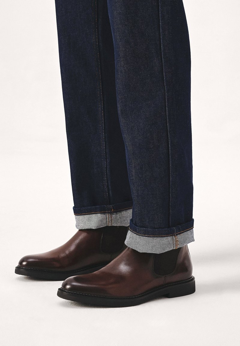 Braune Leder Chelsea-Stiefel mit glatter Oberfläche, runder Zehenpartie und elastischen Seitenpaneelen, kombiniert mit hochgekrempelten dunkelblauen Jeans.