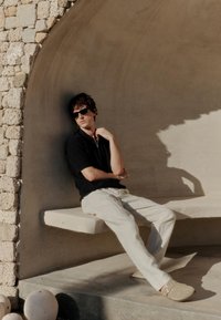 Mlad moški z očali za sonce, črno majico in bež hlačami sedi na kamniti klopi pod obokanim kamnitim zidom na sončni svetlobi.