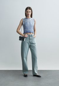 Mladá žena v modrom vzorovanom crop tope bez rukávov, voľných svetlomodrých džínsoch, čiernych balerínkach a s malou čiernou kabelkou cez rameno.